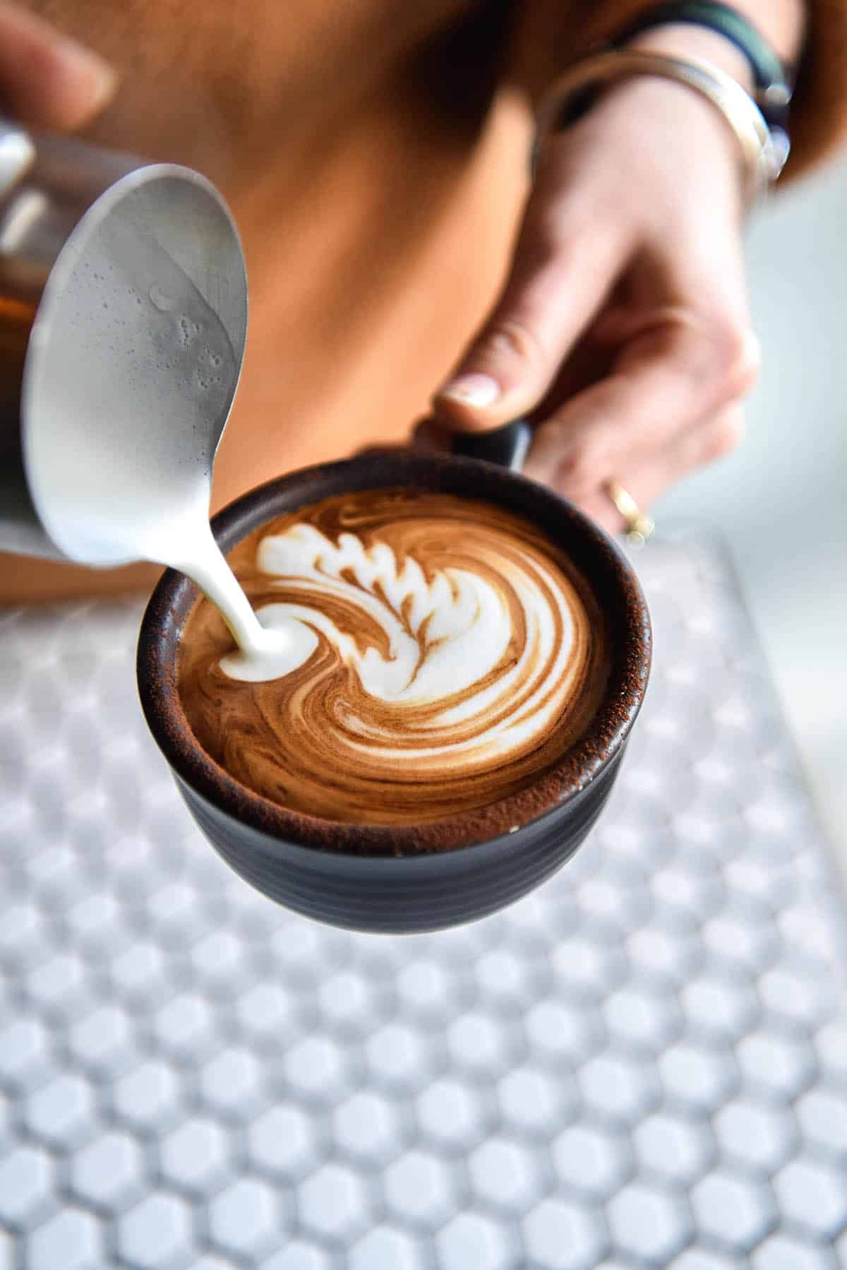 A close up image of a cappuccino being poured into a matte black mug. The cappuccino is being held over a white hexagonal tiled table and the person pouring it wears wings and an orange shirt. 