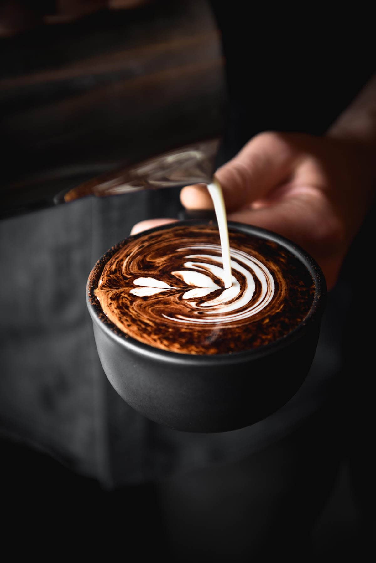 A moody dark image of a cappuccino being poured into a matte black mug. 