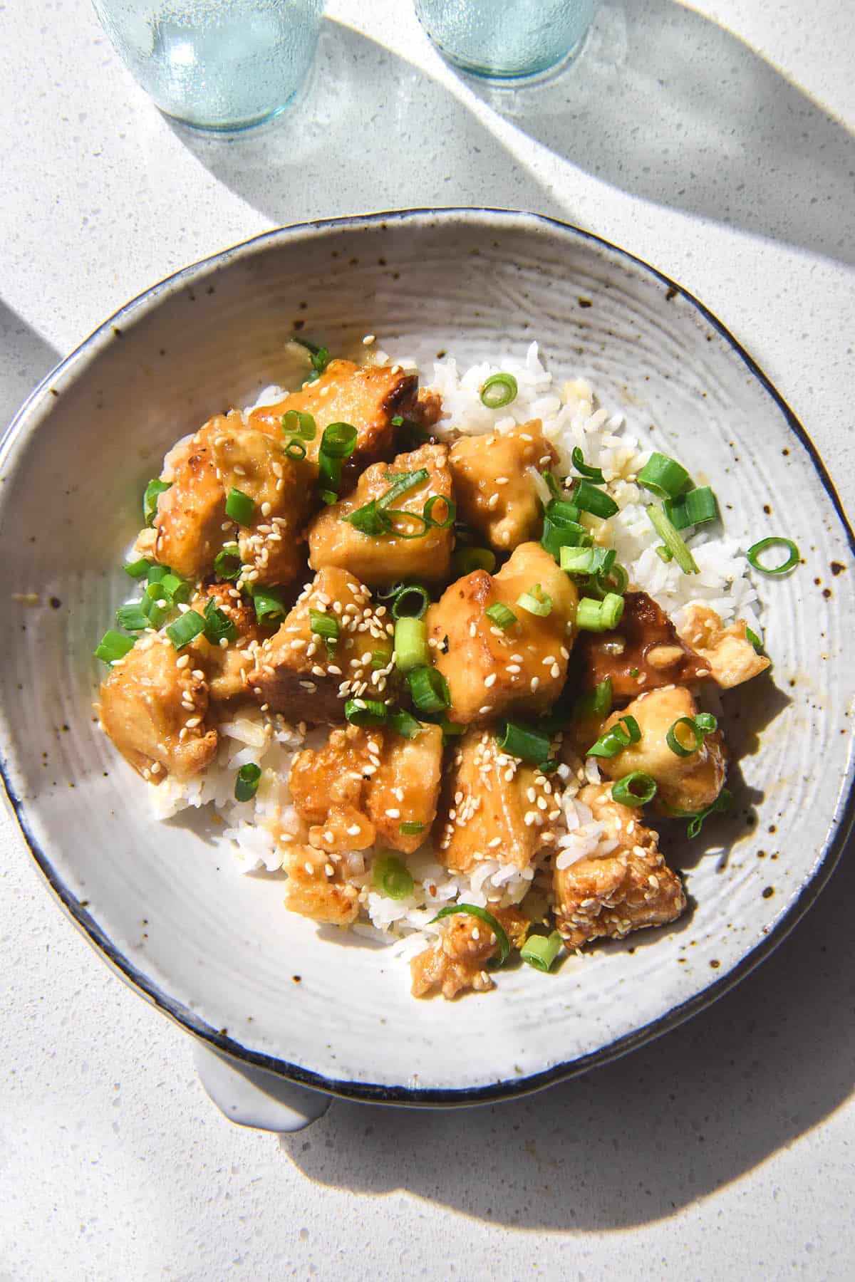 A brightly lit aerial image of a white ceramic bowl filled with lemon tofu, rice, sesame seeds and spring onion greens. The bowl sits atop a white stone benchtop and glasses of water sit in the top of the image. 