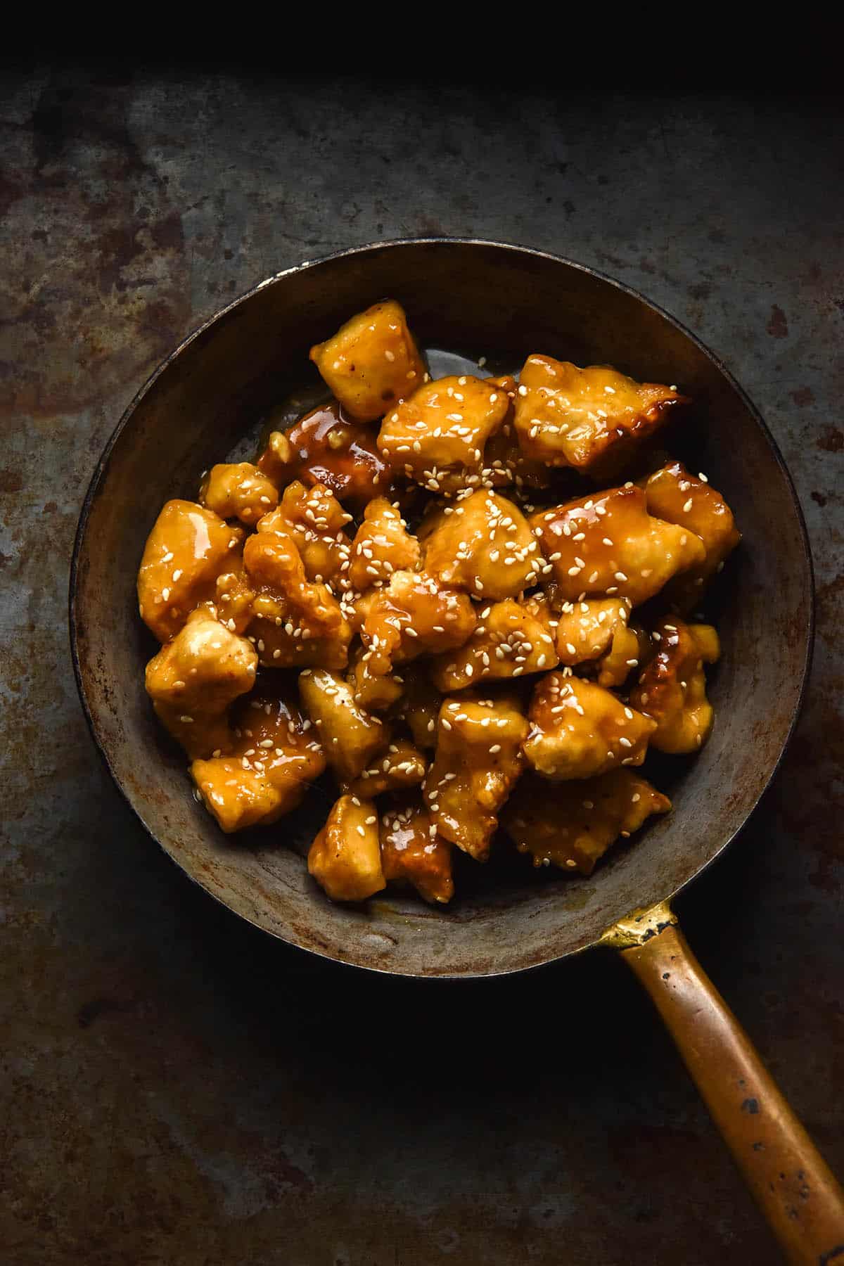An aerial image of a dark steel mini skillet filled with juicy lemon tofu topped with toasted sesame seeds. The skillet sits on a dark steel backdrop.