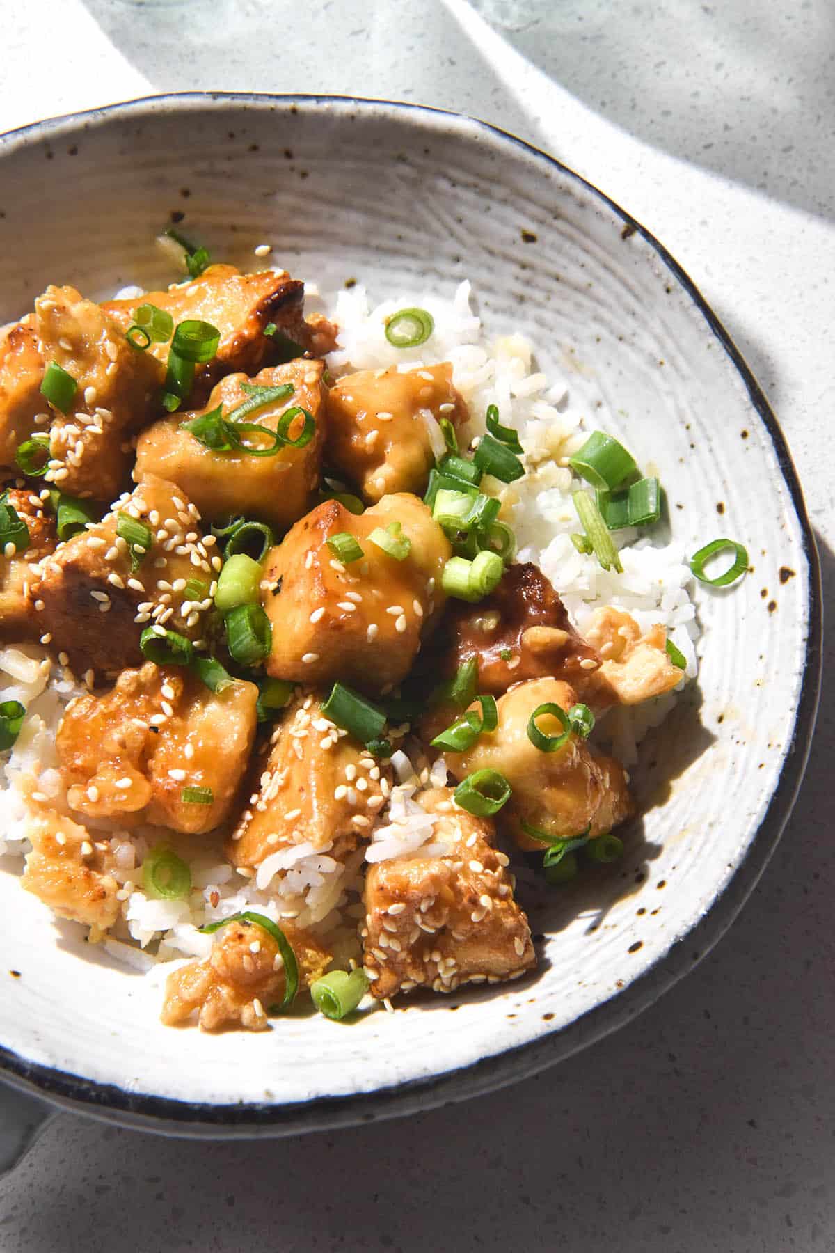 A brightly lit aerial image of a white ceramic bowl filled with lemon tofu, rice, sesame seeds and spring onion greens. The bowl sits atop a white stone benchtop and glasses of water sit in the top of the image. 