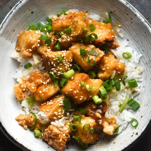 An aerial close up of a white ceramic bowl filled with lemon tofu, rice, sesame seeds and spring onion greens. The bowl sits atop a dark steel backdrop.