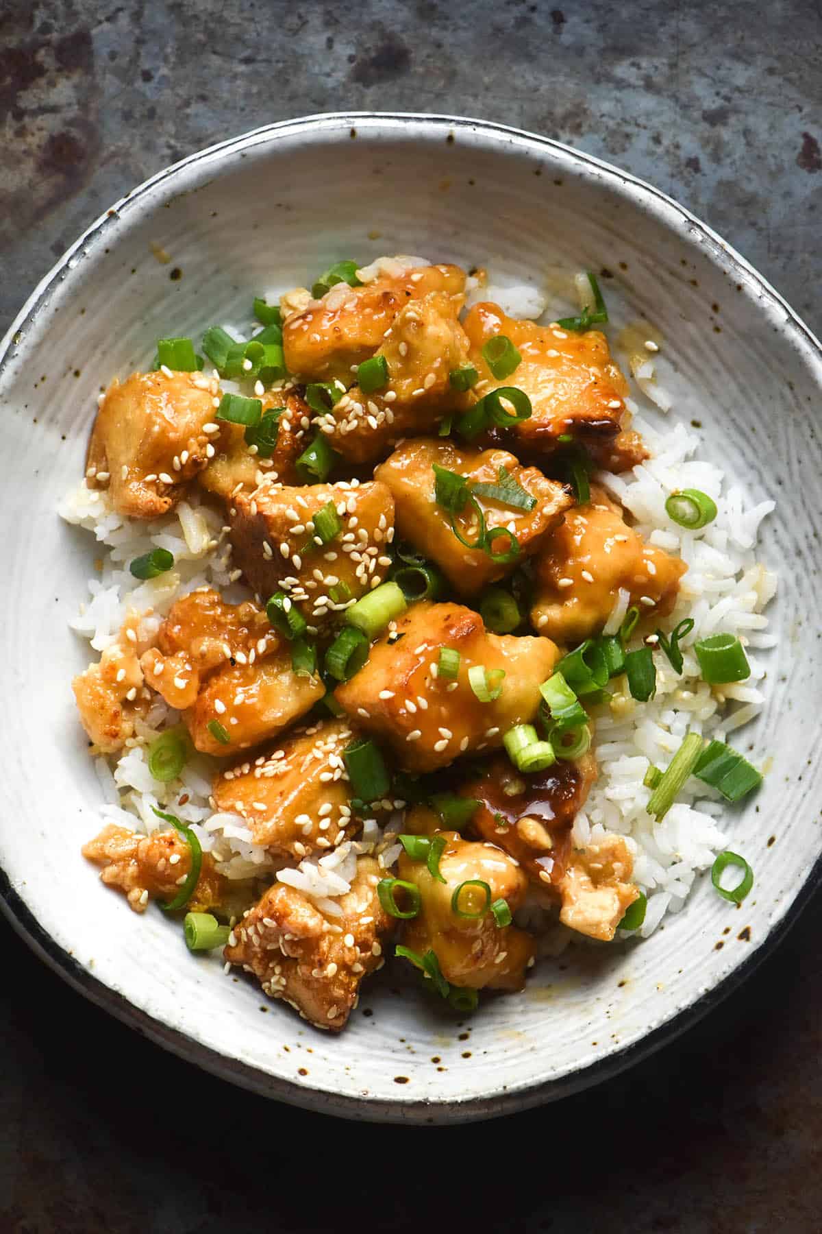 An aerial close up of a white ceramic bowl filled with lemon tofu, rice, sesame seeds and spring onion greens. The bowl sits atop a dark steel backdrop.