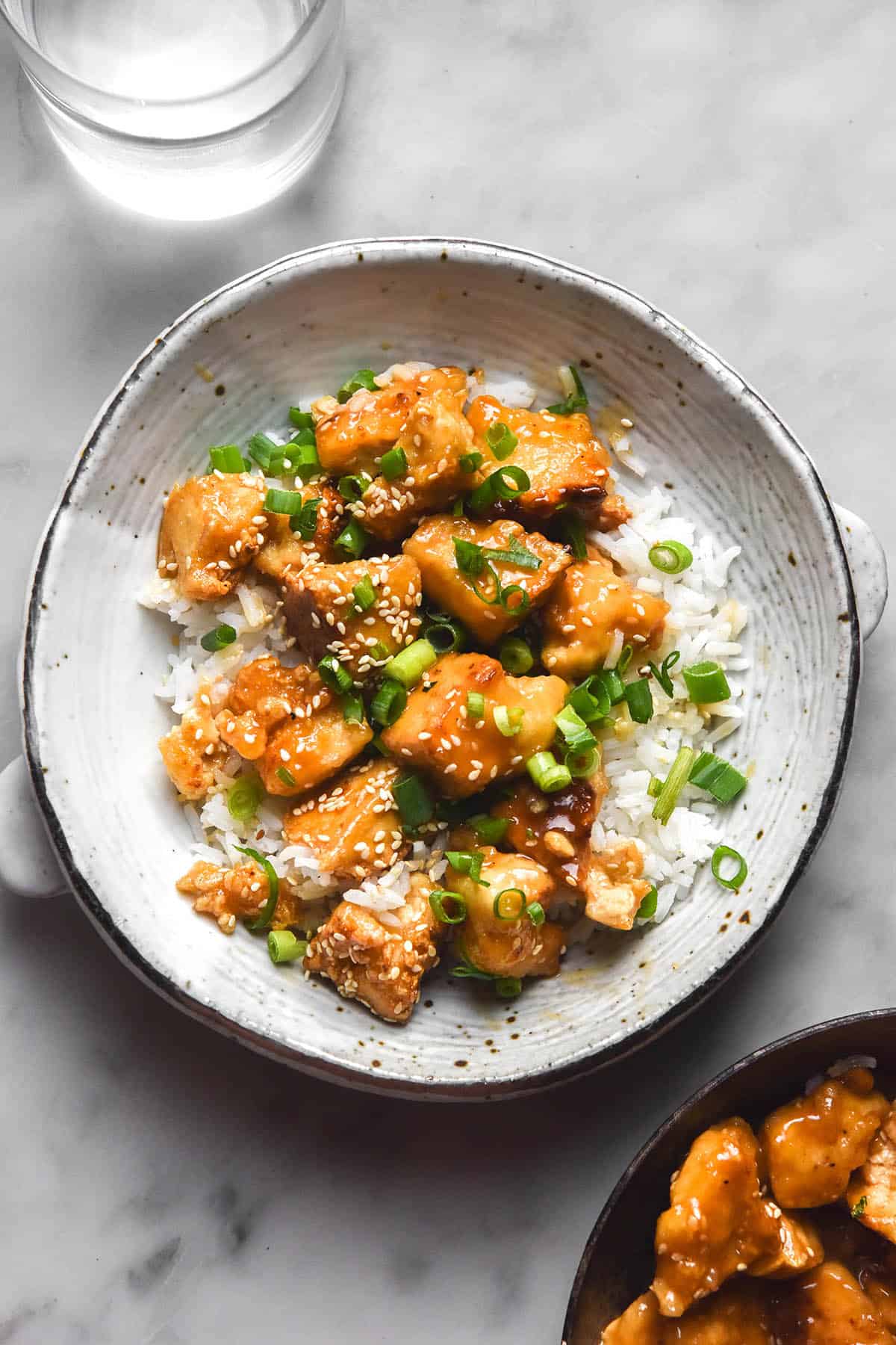 A brightly lit aerial image of a white ceramic bowl filled with lemon tofu, rice, sesame seeds and spring onion greens. The bowl sits atop a white marble table and glasses of water sit in the top of the image. A skillet of extra lemon tofu sits in the bottom right of the image. 