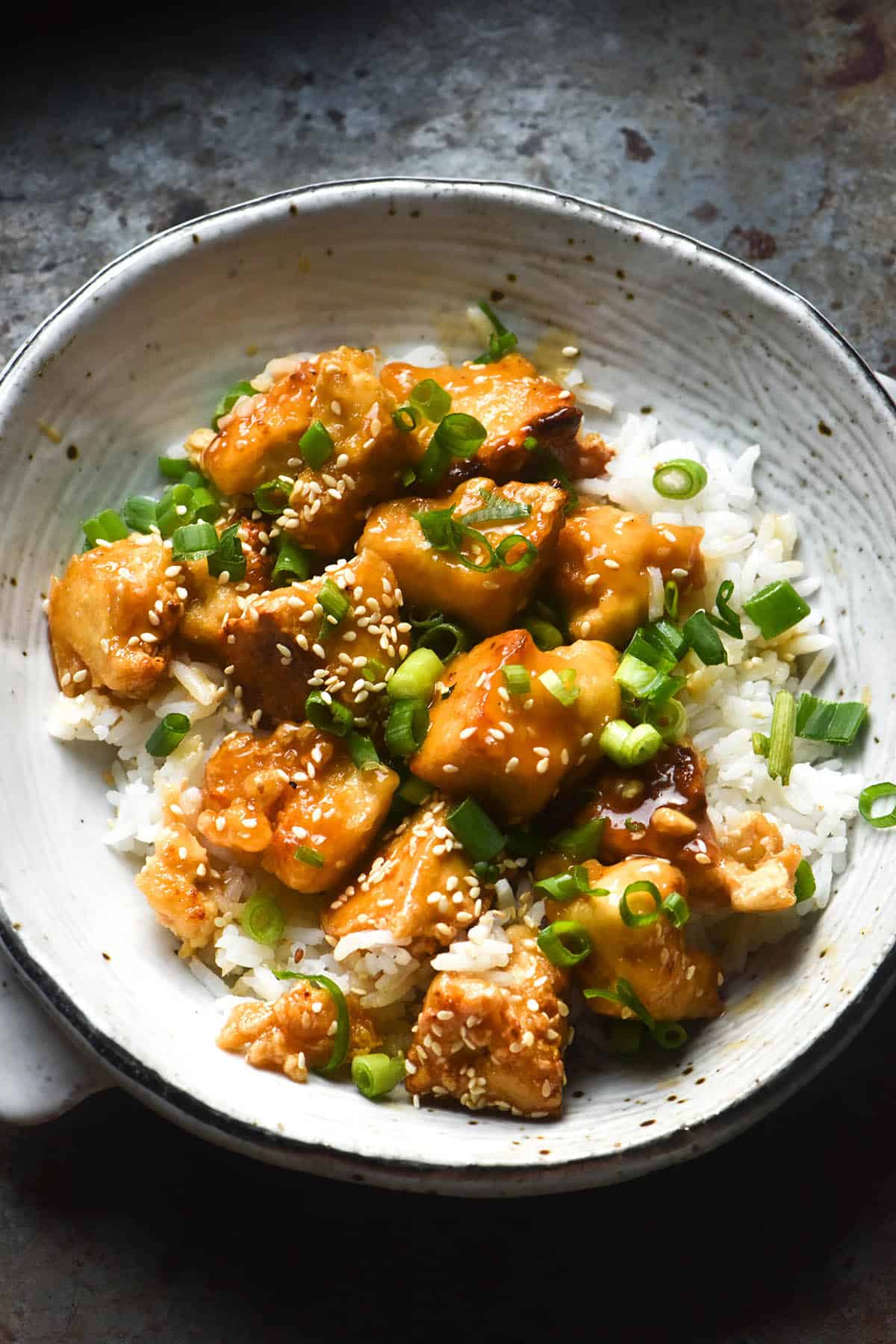 A close up aerial image of a white ceramic bowl filled with lemon tofu, white rice, sesame seeds and spring onion greens. The bowl sits on a dark ceramic backdrop.