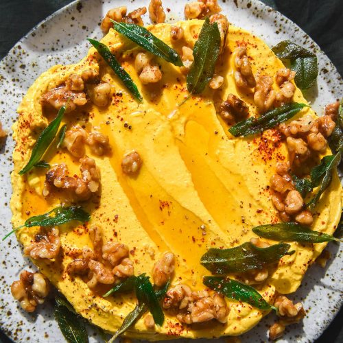 An aerial close up image of a white speckled ceramic plate topped with pumpkin dip. The dip is adorned with candied walnuts and crispy sage leaves. The plate sits on a dark olive linen backdrop and two glasses of water sit to the top of the image.