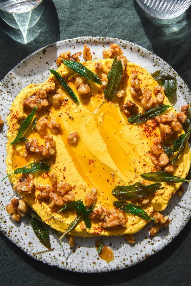 An aerial close up image of a white speckled ceramic plate topped with pumpkin dip. The dip is adorned with candied walnuts and crispy sage leaves. The plate sits on a dark olive linen backdrop and two glasses of water sit to the top of the image.