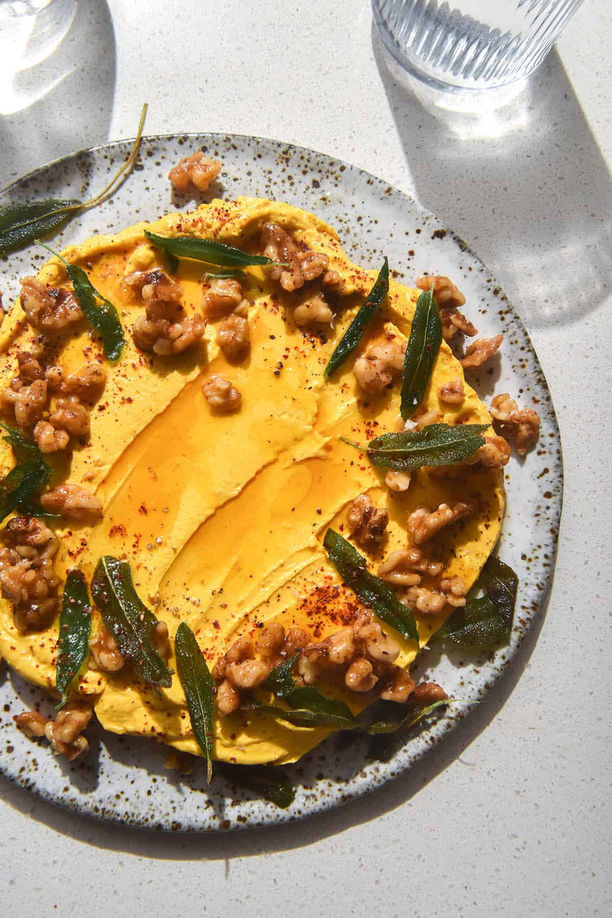 An aerial image of a white speckled ceramic plate topped with low FODMAP pumpkin feta dip. The dip is adorned with a wreath of candied walnuts and crispy sage leaves. The plate sits atop a white stone benchtop and glasses of water sit to the top of the image.