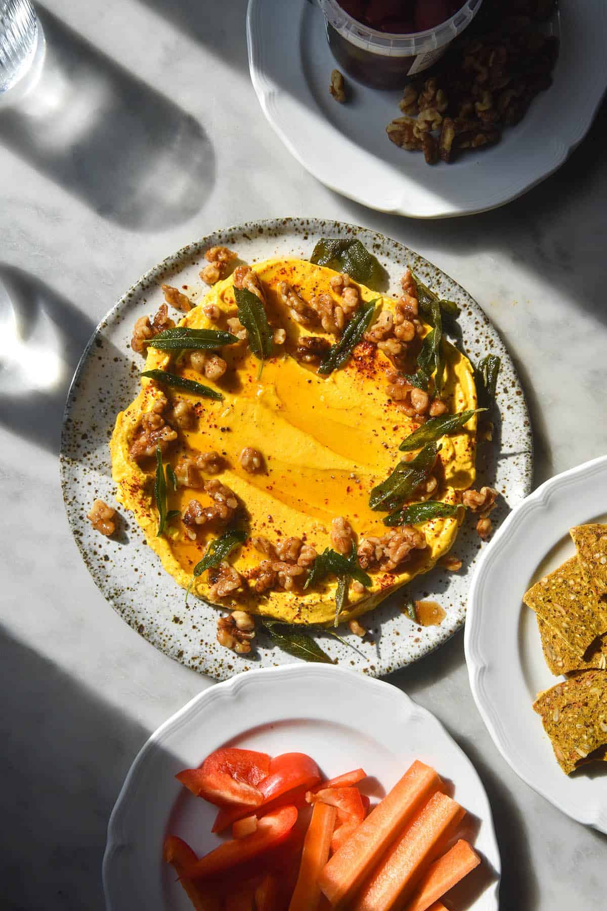 An aerial image of a white speckled ceramic plate topped with pumpkin dip on a white marble table. The plate of dip is adorned with a wreath of candied walnuts and crispy sage leaves. The plate is surrounded by other smaller white plates of crackers, nuts and crudites. 