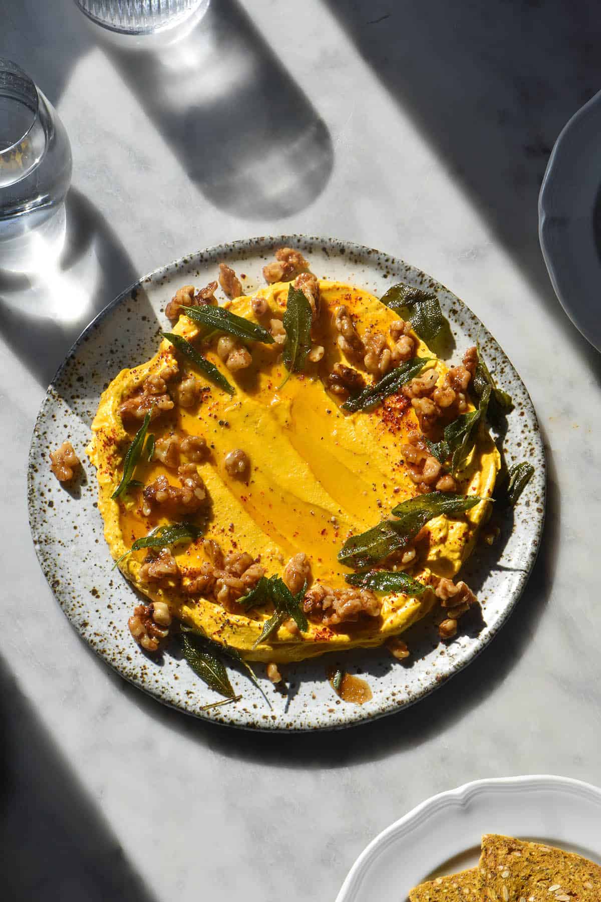 An aerial image of a plate of pumpkin feta dip on a marble table in contrasting sunlight. A water glass sits to the top left of the image and plates are casually arranged around the edges. 