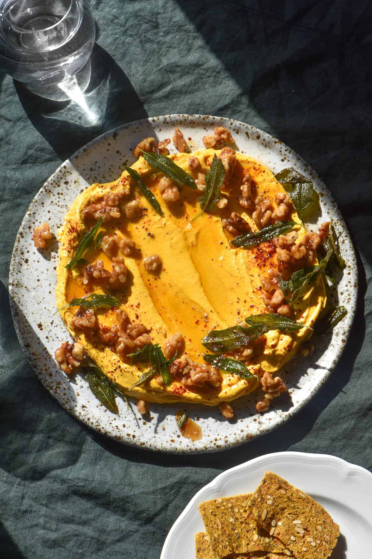An aerial sunlit image of a white ceramic plate topped with pumpkin feta dip. The dip is decorated with candied walnuts, crispy sage leaves and chilli flakes. It sits atop a dark olive linen tablecloth and a water glass sits to the top left of the image. A white ceramic plate of crackers sits to the bottom right of the dip. 