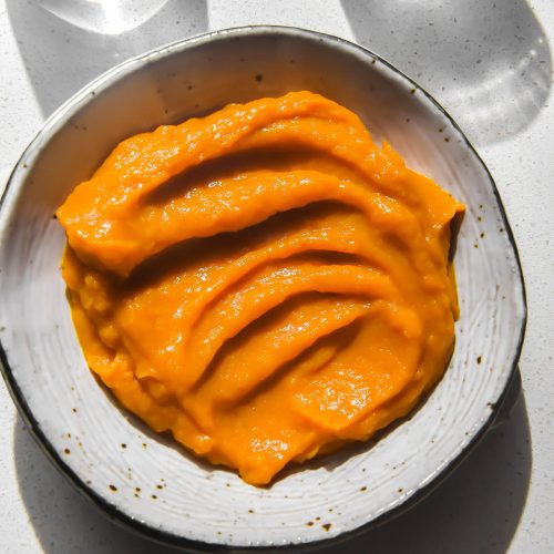 An aerial image of a white ceramic bowl filled with microwave pumpkin puree on a white stone benchtop. The benchtop is in bright light and two water glasses sit to the top of the puree.