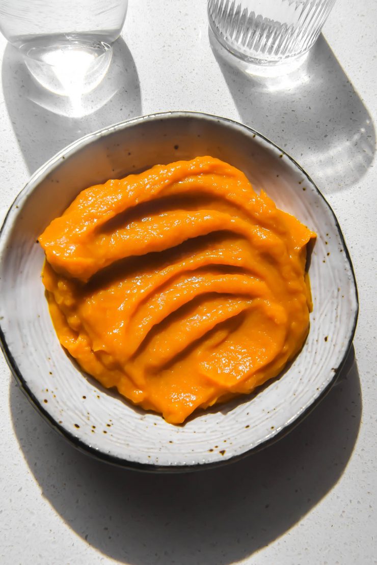 An aerial image of a white ceramic bowl filled with microwave pumpkin puree on a white stone benchtop. The benchtop is in bright light and two water glasses sit to the top of the puree.