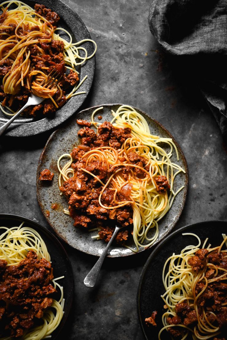 An aerial image of three dark blue plates of vegan tofu spaghetti bolognese on a mottled dark blue steel backdrop.