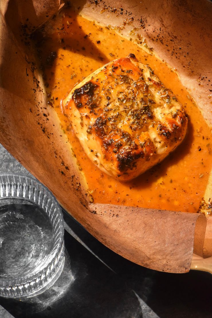 An aerial image of baked haloumi on a baking sheet in a ceramic baking dish. The dish sits on a dark backdrop in bright sunlight, and a sunlit glass of water sits to the left of the haloumi.