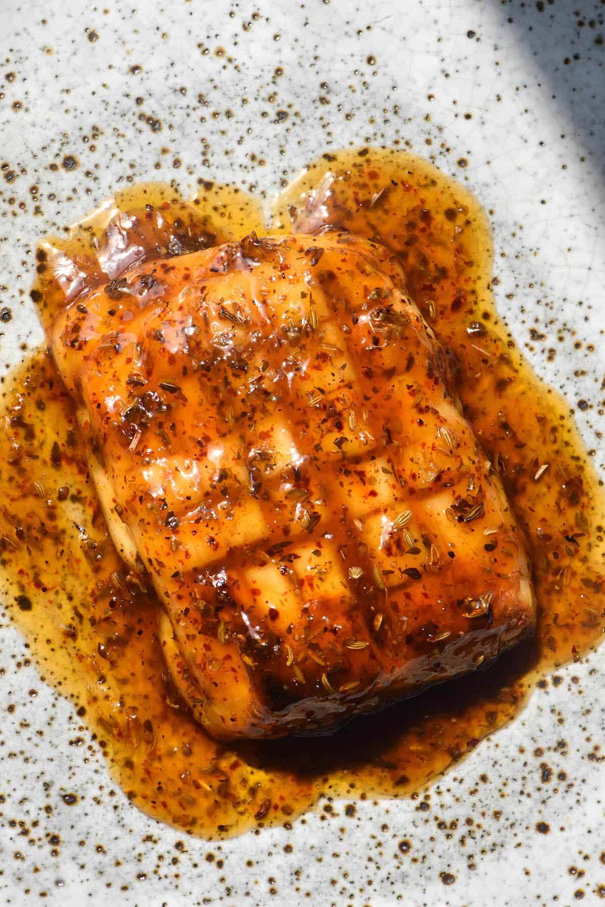 An aerial sunlit image of a slab of baked halloumi that has been scored and smothered in a maple and lemon glaze. The glaze is a deep red and is dotted with fennel seeds, dried oregano and chilli flakes. The halloumi sits atop a white speckled ceramic plate. 