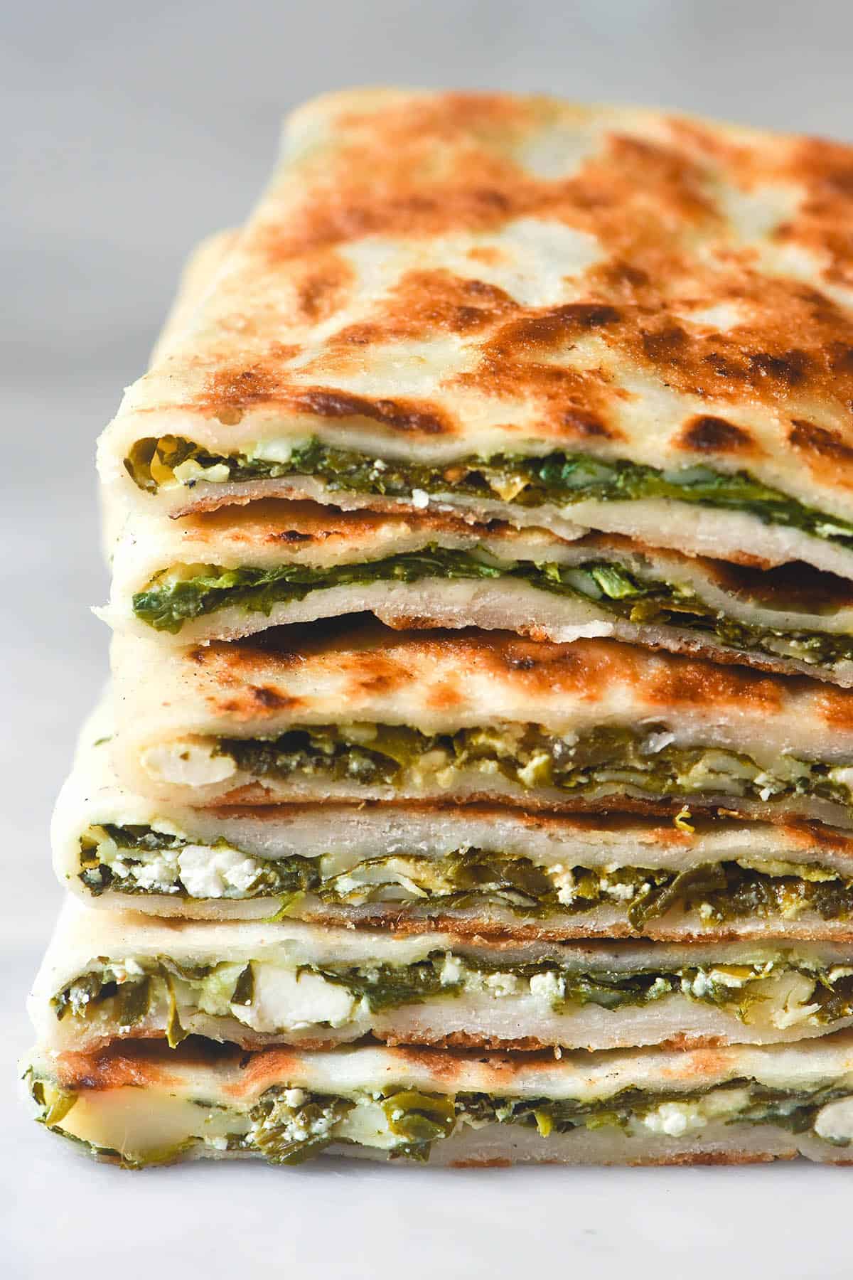 A macro image of a stack of sliced gluten free spinach and feta gozleme on a white marble table against a white backdrop. The sliced section of the gozleme faces the camera, revealing the spinach and feta filling. 