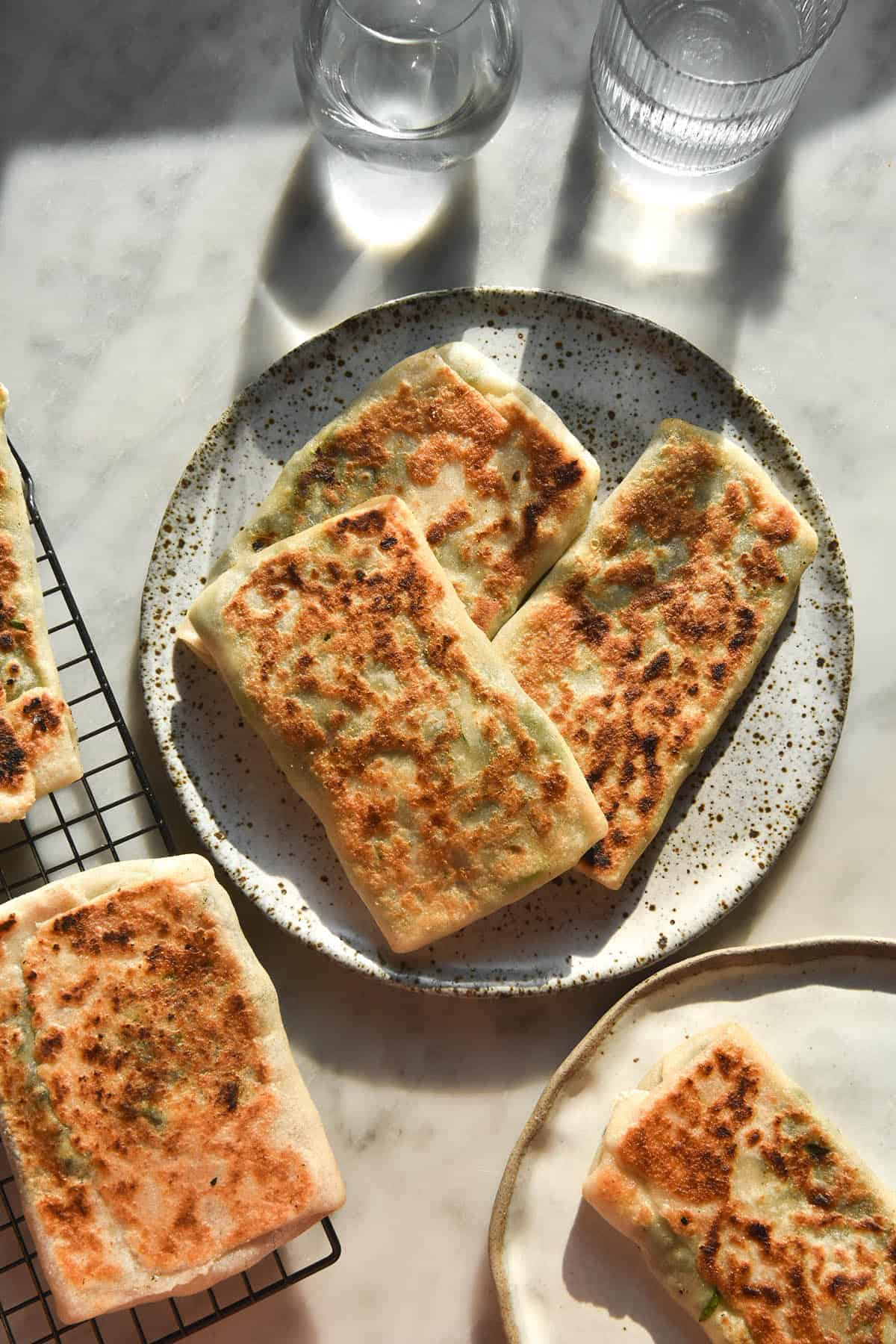An aerial image of gluten free gozleme on a white speckled ceramic plate and cooling rack atop a white marble table in contrasting sunlight. 
