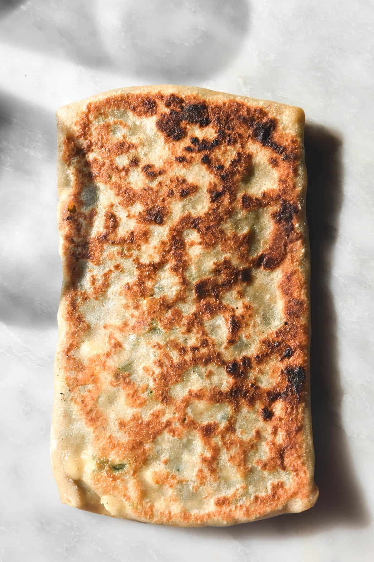 An aerial macro image of a deeply golden gluten free gozleme atop a sunlit white marble table 