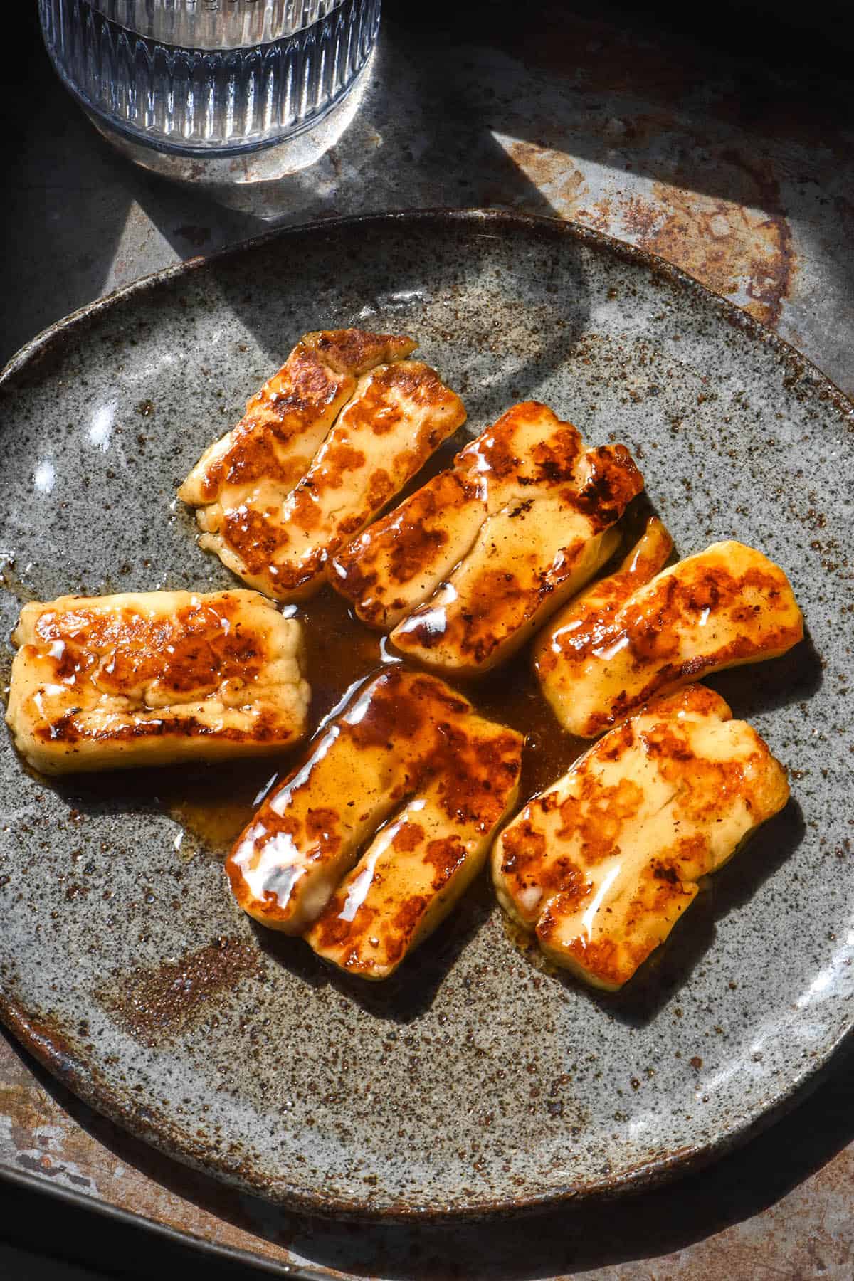 An aerial image of golden brown halloumi pieces with a honey lemon glaze. The halloumi sits atop a dark speckled ceramic plate on a dark steel backdrop