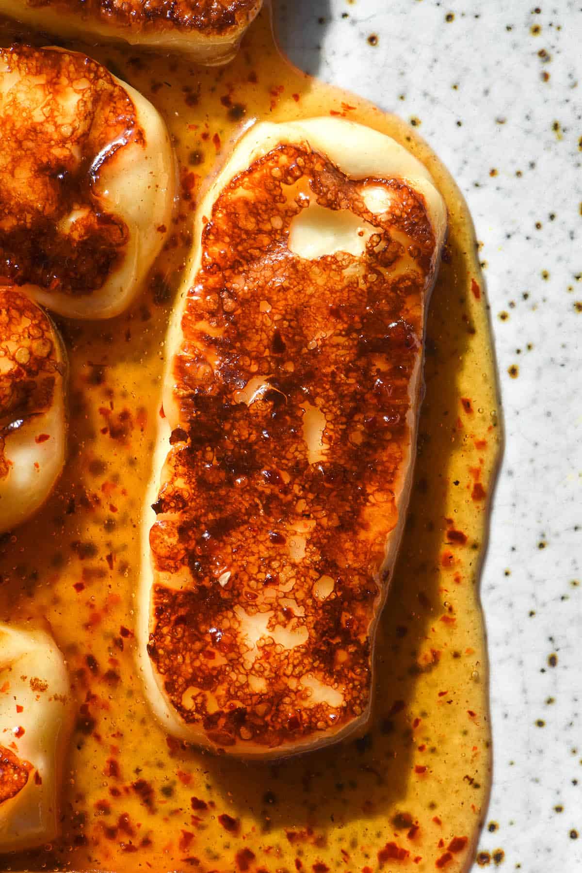 An aerial macro image of golden brown honey halloumi on a white speckled ceramic plate. The sauce is golden brown and flecked with red chilli flakes