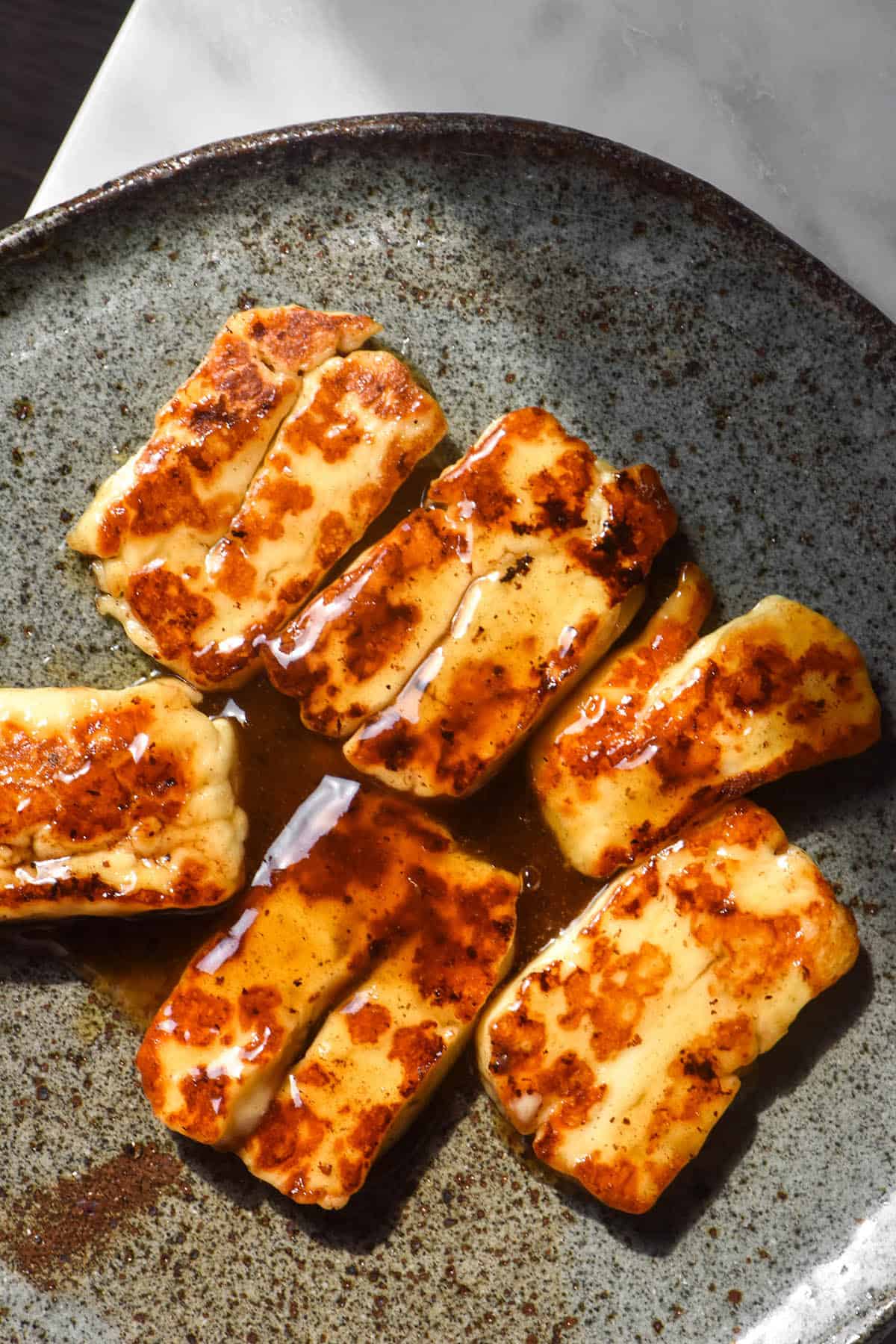 An aerial image of golden brown halloumi pieces with a honey lemon glaze. The halloumi sits atop a dark speckled ceramic plate on a white marble table. 