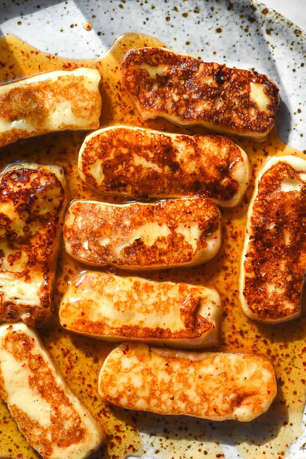 An aerial image of golden brown honey glazed haloumi on a white speckled ceramic plate in bright sunlight. 