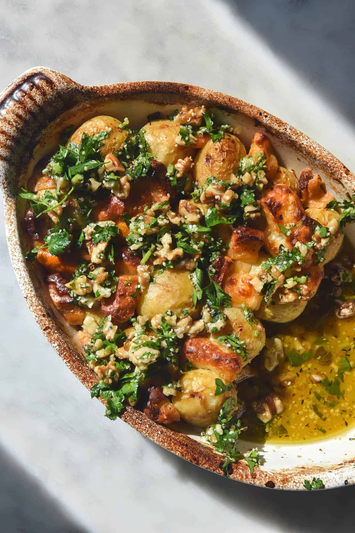 An aerial image of lemon roasted potatoes baked with halloumi and finished with a herb and walnut salsa. The potatoes sit in a ceramic baking dish top a white marble table in bright sunlight. 