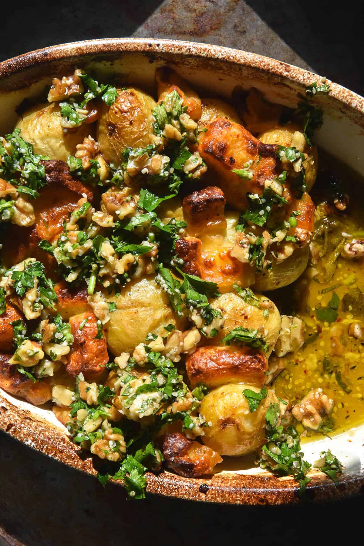 An aerial close up image of a baking dish filled with lemon baked potatoes topped with haloumi and a walnut herb salsa. The dish sits atop a dark steel backdrop in bright sunlight. 