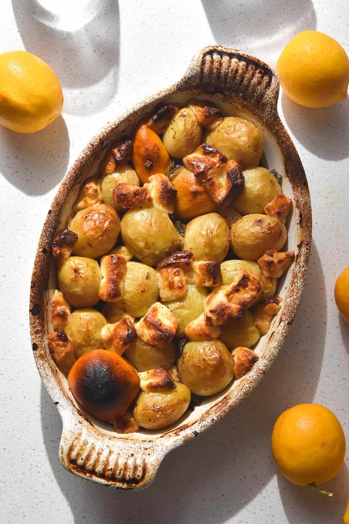 An aerial image of a lemon potato and haloumi bake in a ceramic dish atop a white stone bench top. The bench is in bright sunlight and is surrounded by lemons. 