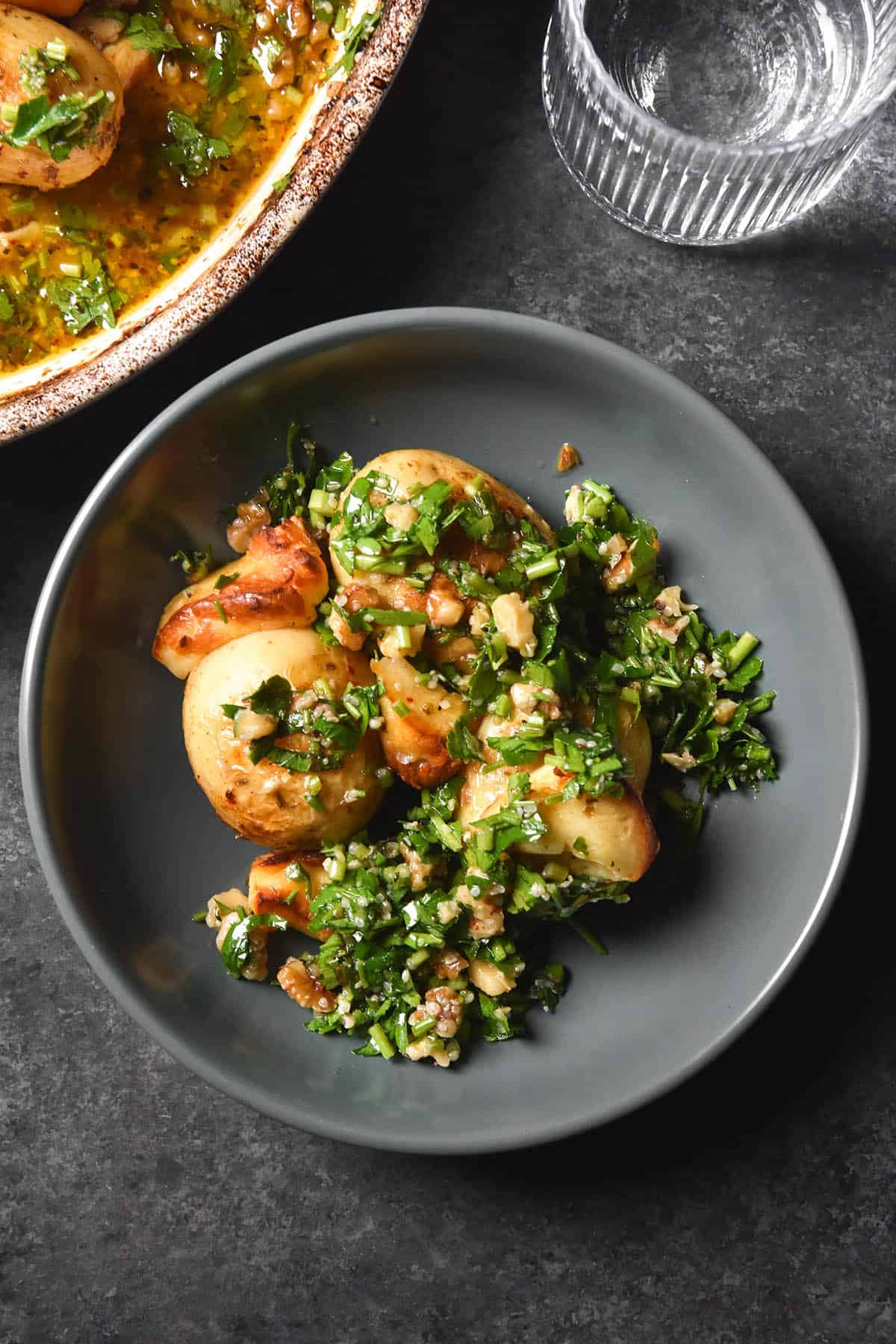 An aerial image of a dark grey ceramic bowl filled with a lemon roasted potato and haloumi bake topped with a walnut and herb salsa. The bowl sits on a dark grey backdrop and a glass of water sits to the top right of the image while the tray bake sits to the top left. 