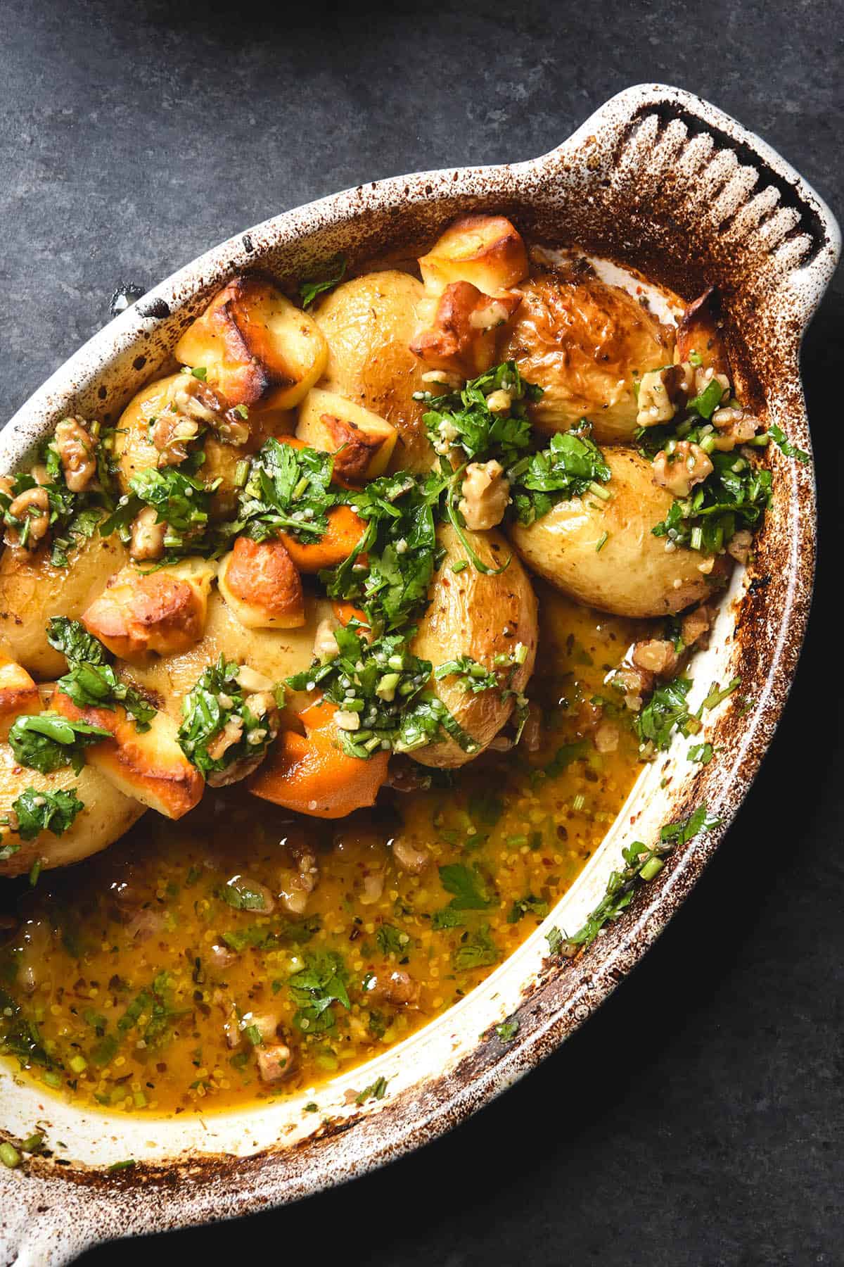 An aerial image of lemon baked potatoes with haloumi and a walnut herb salsa in a white ceramic baking dish atop a dark grey backdrop.