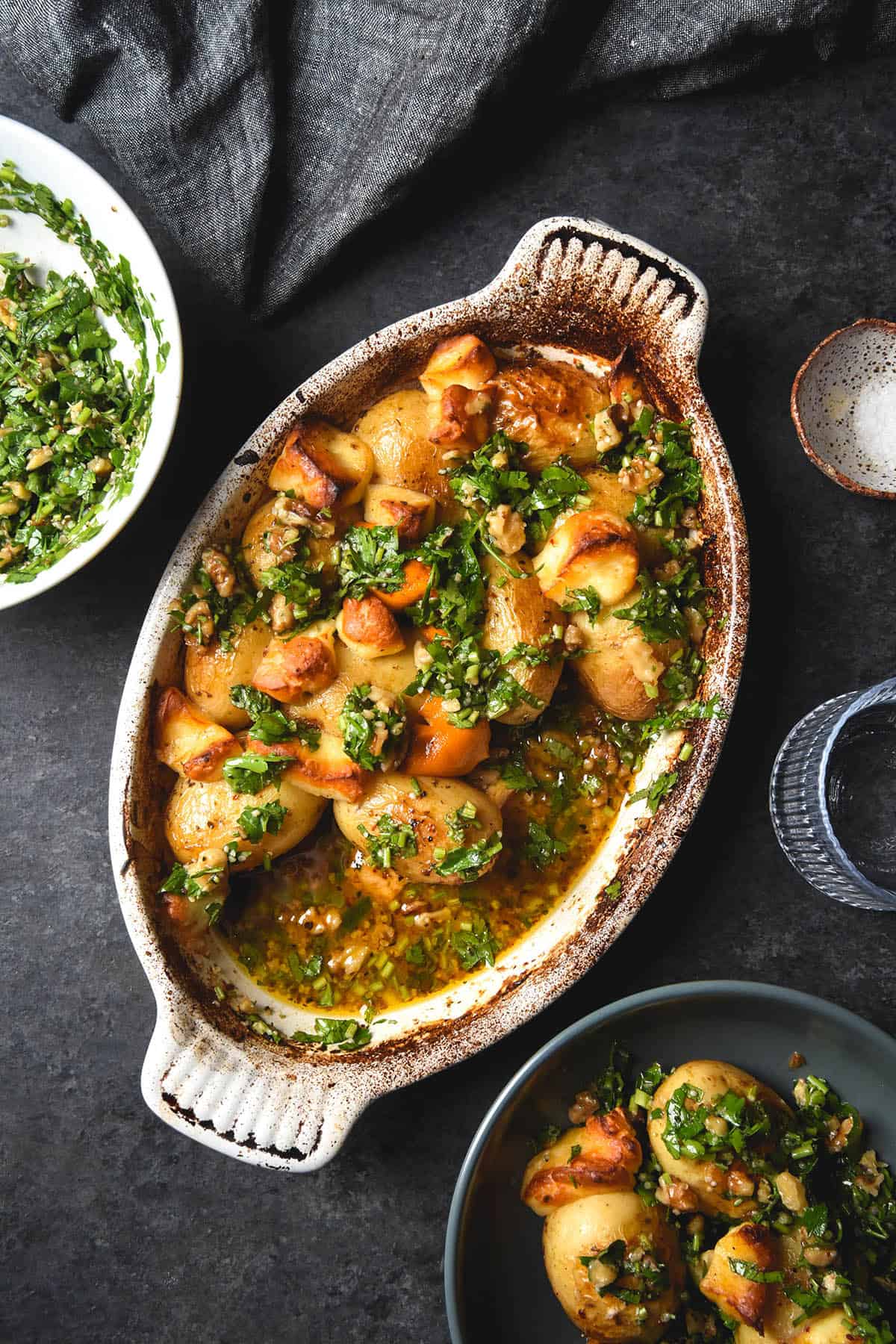 An aerial image of lemon baked potatoes with haloumi and a walnut herb salsa in a white ceramic baking dish atop a dark grey backdrop. The central dish is surrounded by extra bowls of the potatoes, water glasses and a bowl of the walnut salsa. 