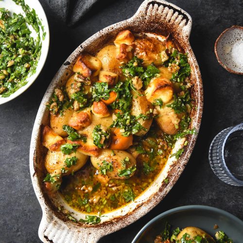 Lemon roasted potatoes with haloumi and a herb salsa in a ceramic baking dish atop a dark blue backdrop. The dish is surrounded by a plate of the potatoes, water glasses and extra herb salsa