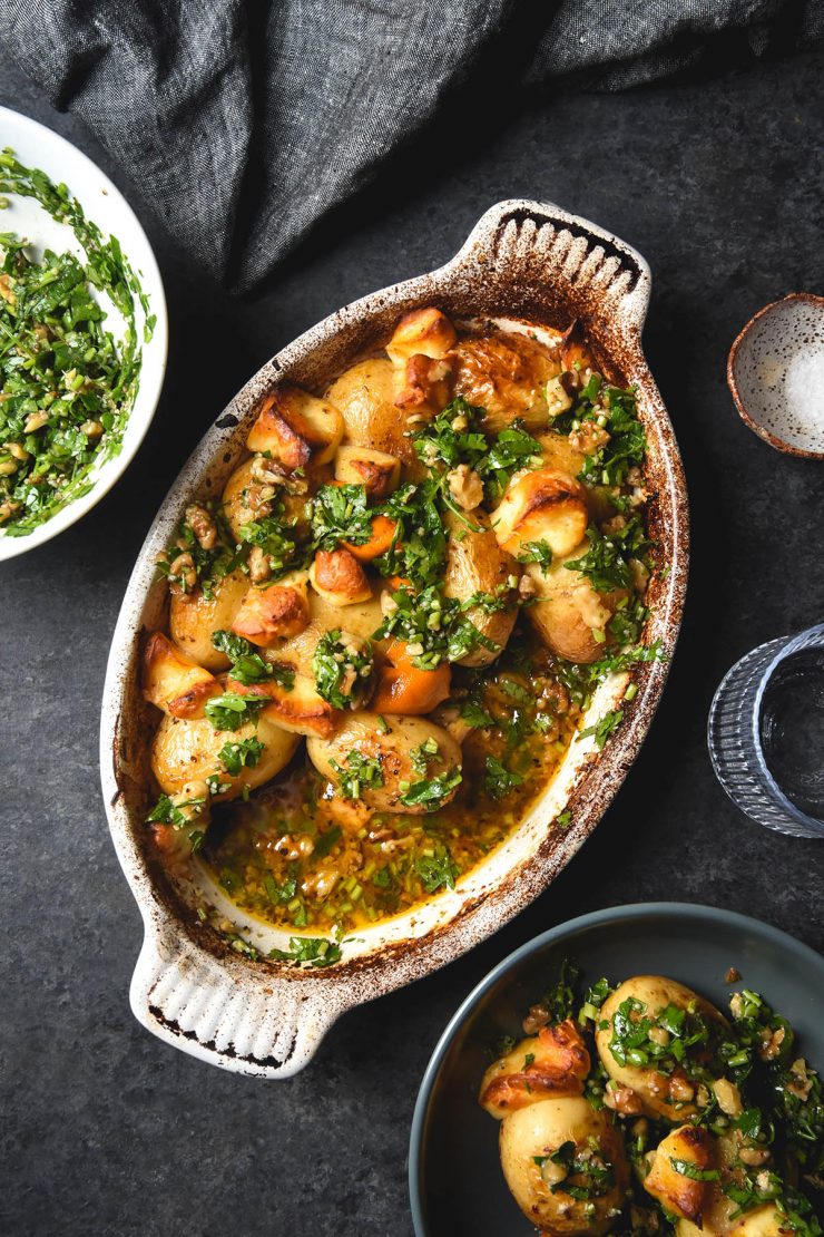 Lemon roasted potatoes with haloumi and a herb salsa in a ceramic baking dish atop a dark blue backdrop. The dish is surrounded by a plate of the potatoes, water glasses and extra herb salsa