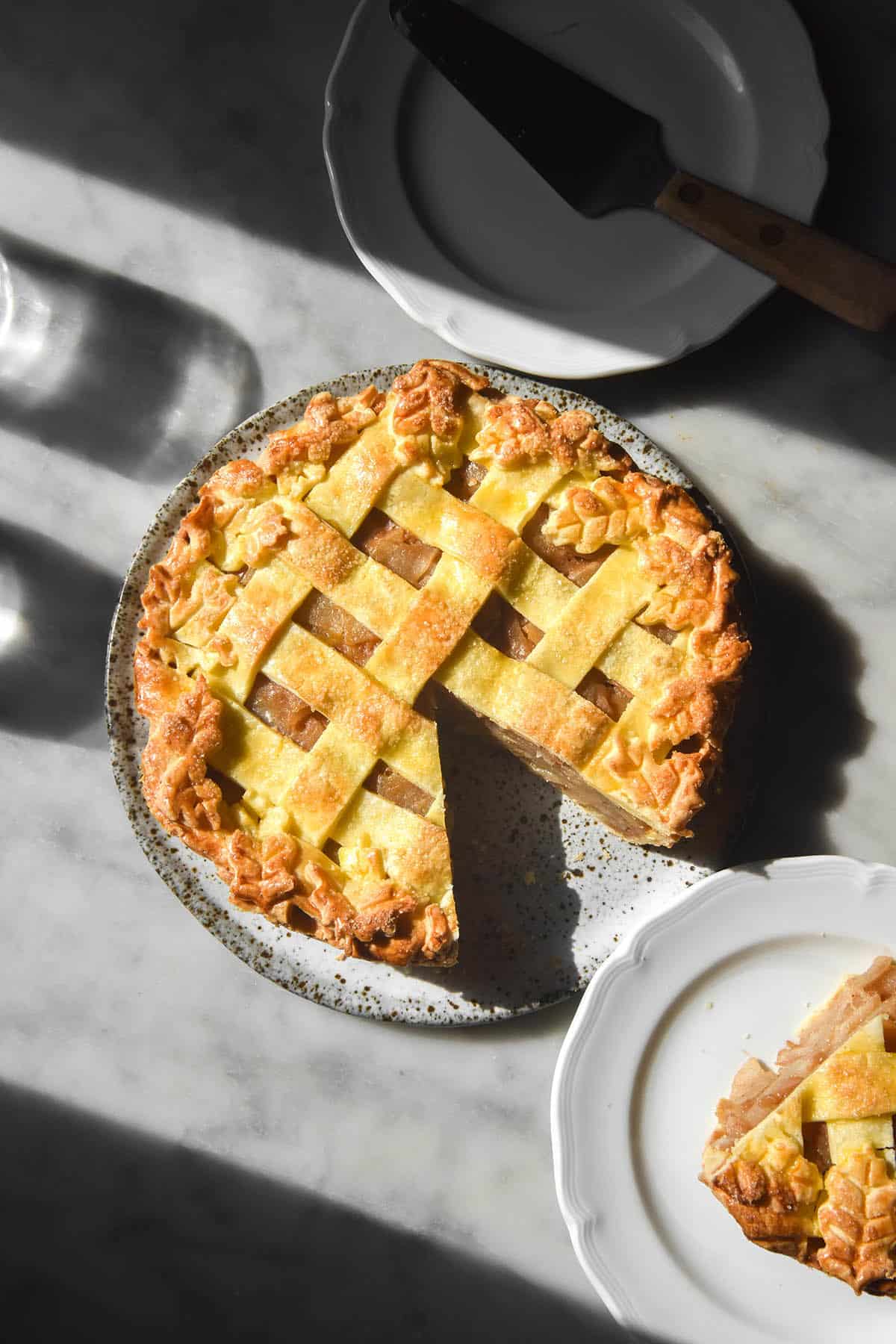 A sunlit aerial image of a gluten free low FODMAP apple pie on a white speckled ceramic plate atop a white marble table. The pie has a lattice top and the edges are decorated with pastry leave. A slice has been removed and sits on a white scalloped plate the bottom right of the image.