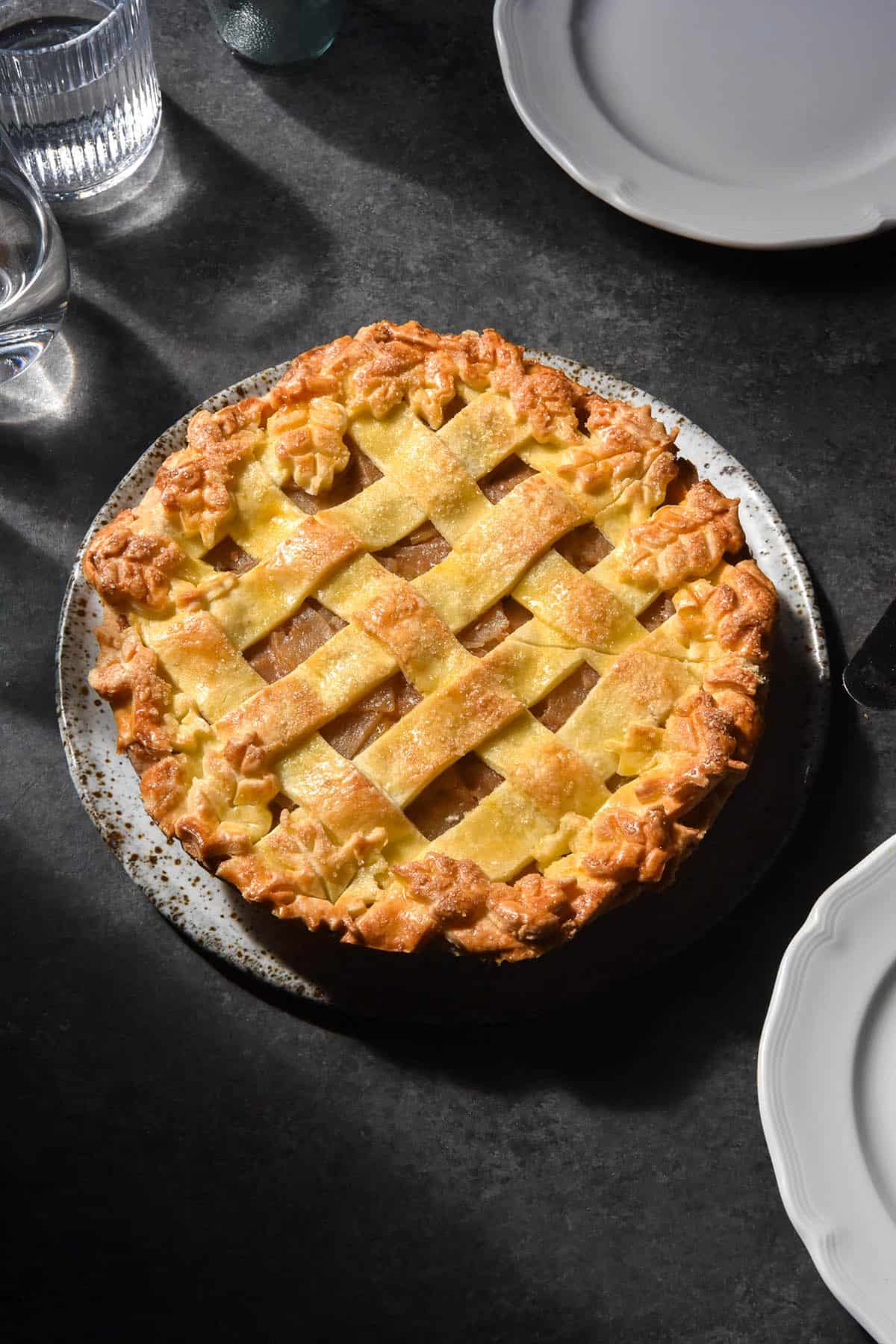 A dark and moody aerial image of a low FODMAP apple pie with a lattice top and pastry leaf decor around the edges. The pie sits on a white speckled ceramic plate atop a dark backdrop. It is surrounded by plates and sunlit glasses of water.