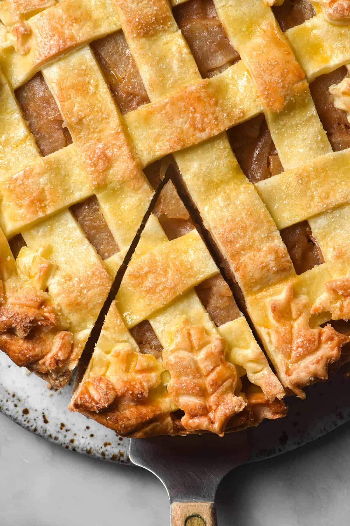 An aerial close up image of a gluten free low FODMAP apple pie. The pie has a lattice top with pastry leaf decor around the edges. It sits on a white ceramic plate atop a white marble table. The pie has been sliced and a cake server with a wooden handle sits underneath the slice.