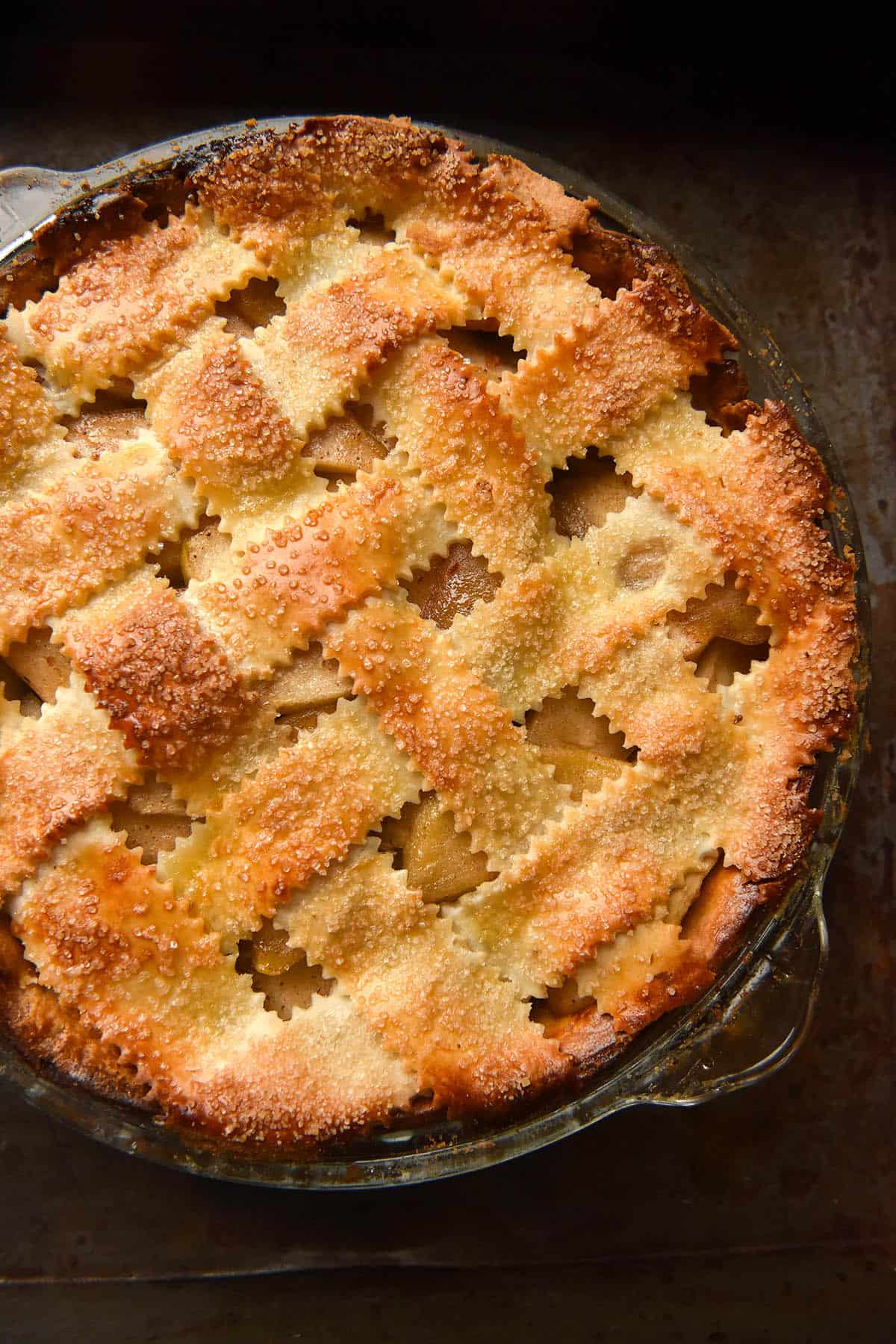 An aerial image of a low FODMAP apple pie with a golden brown lattice topping topped with sugar. The pie sits in a glass pie dish atop a dark steel backdrop.