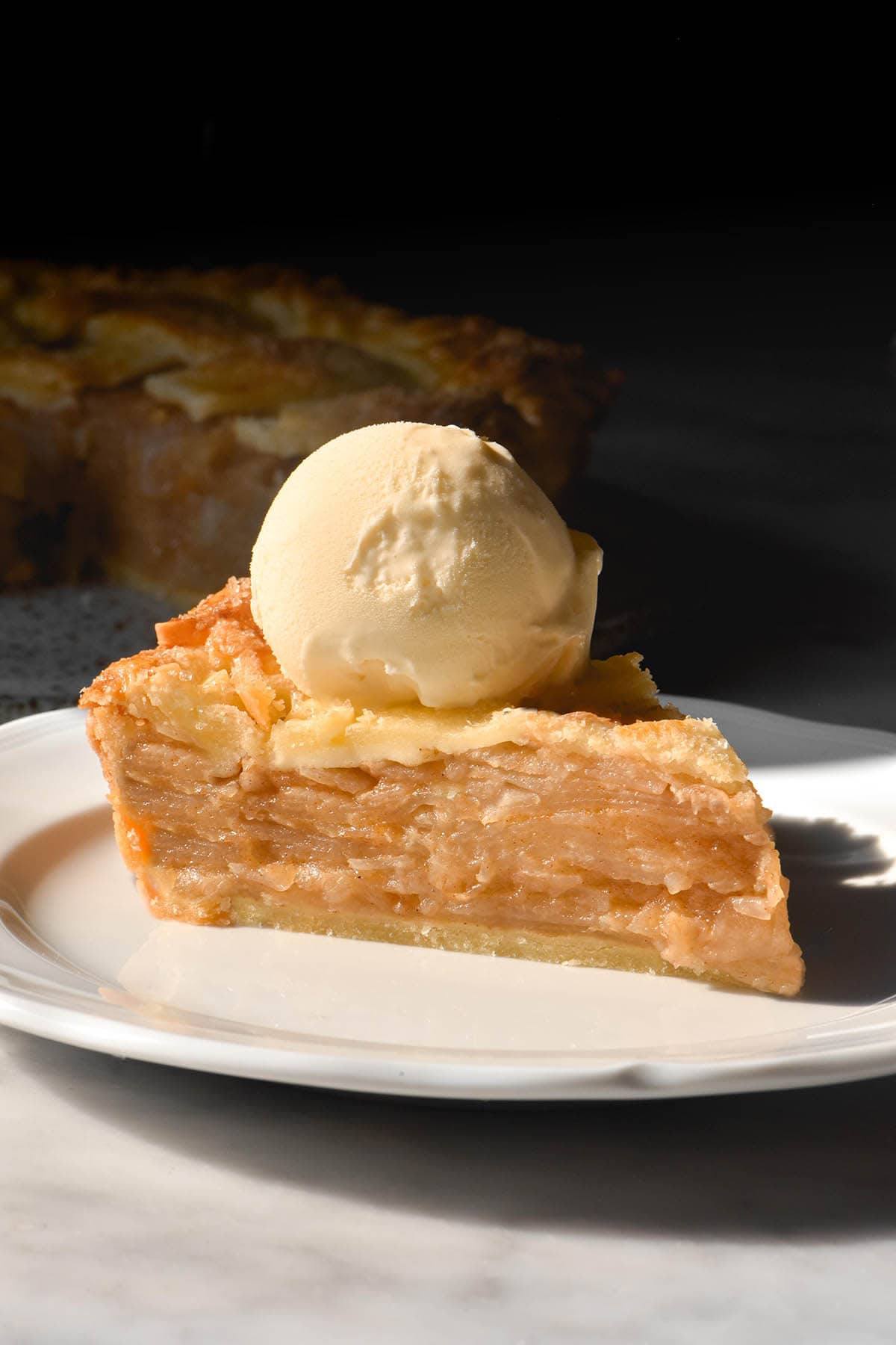 A side on image of a slice of low FODMAP apple pie on a white scalloped plate topped with a scoop of vanilla ice cream. The plate sits atop a white marble table against a dark backdrop.