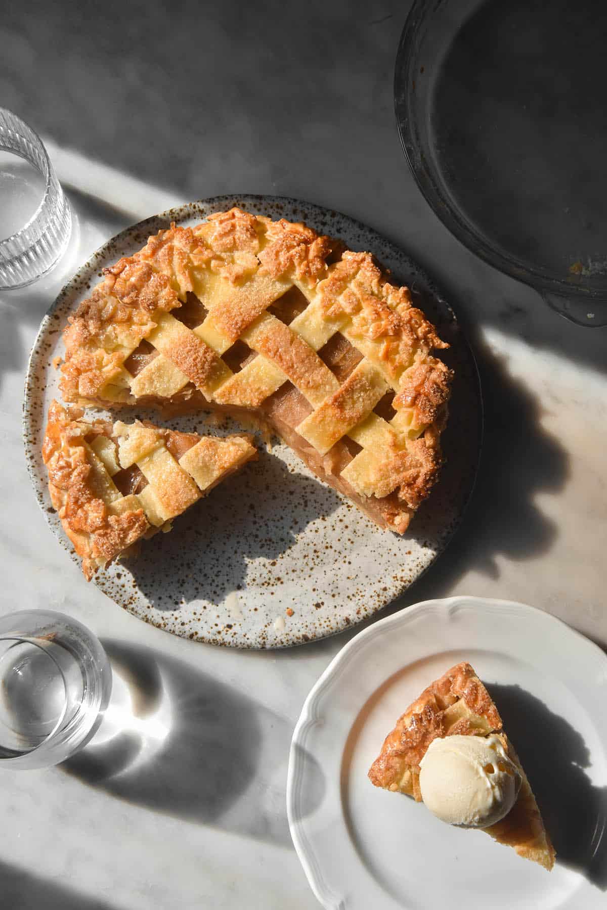 An aerial image of a low FODMAP apple pie atop a white speckled ceramic plate on a white marble table in contrasting sunlight. The pie top is golden brown lattice top and a slice of pie topped with vanilla ice cream sits to the bottom right of the image.
