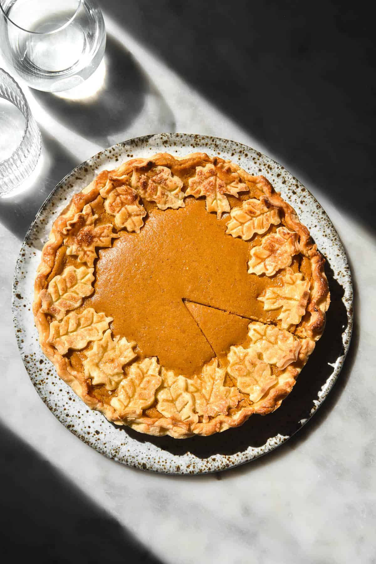 An aerial image of a low FODMAP pumpkin pie on a white marble table in contrasting sunlight. The pie sits on a white speckled ceramic plate and is decorated with a ring of golden brown gluten free pastry leaves. 