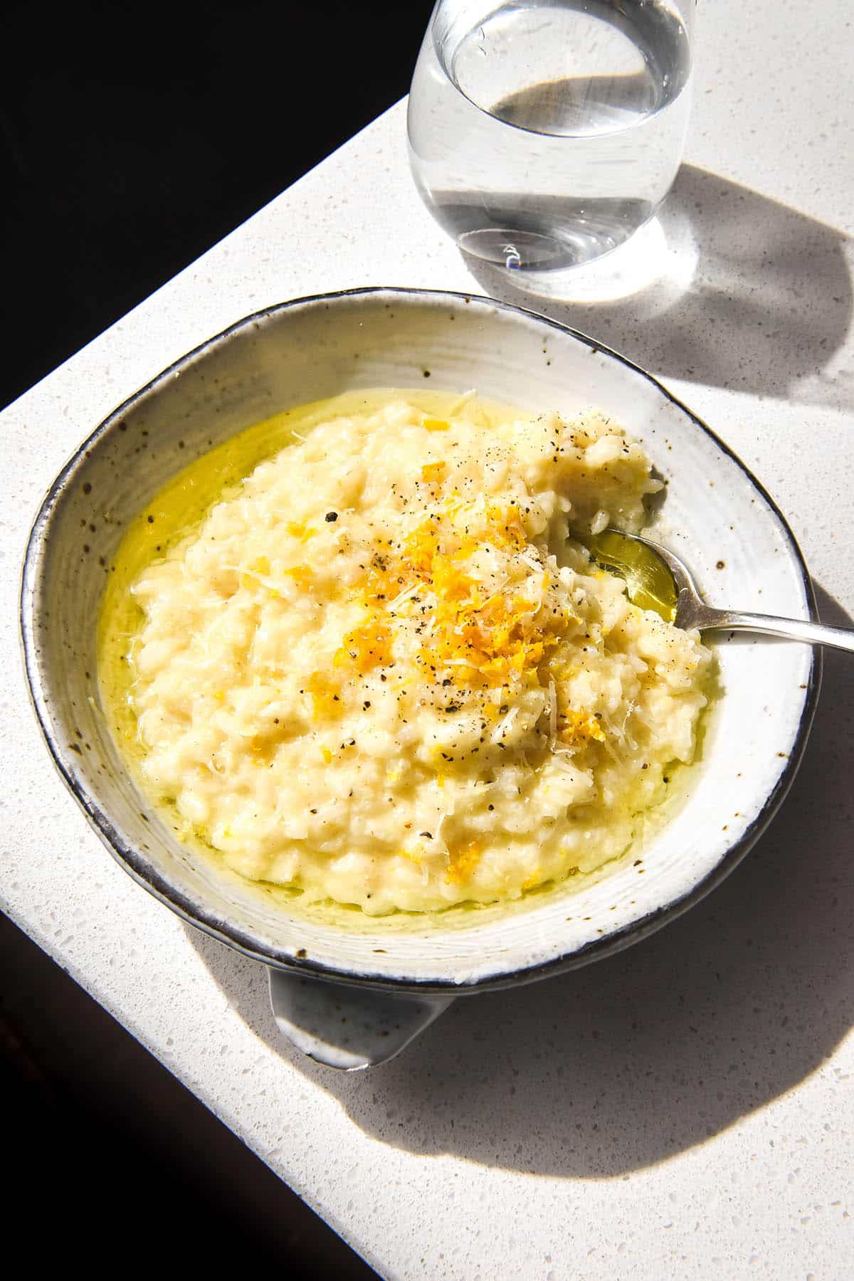 A brightly lit aerial image of a white ceramic bowl filled with low FODMAP risotto al limone. The risotto is topped with garlic infused oil, grated parmesan, lemon zest and black pepper. It sits atop a white stone benchtop and a sunlit glass of water sits to the top right of the image.