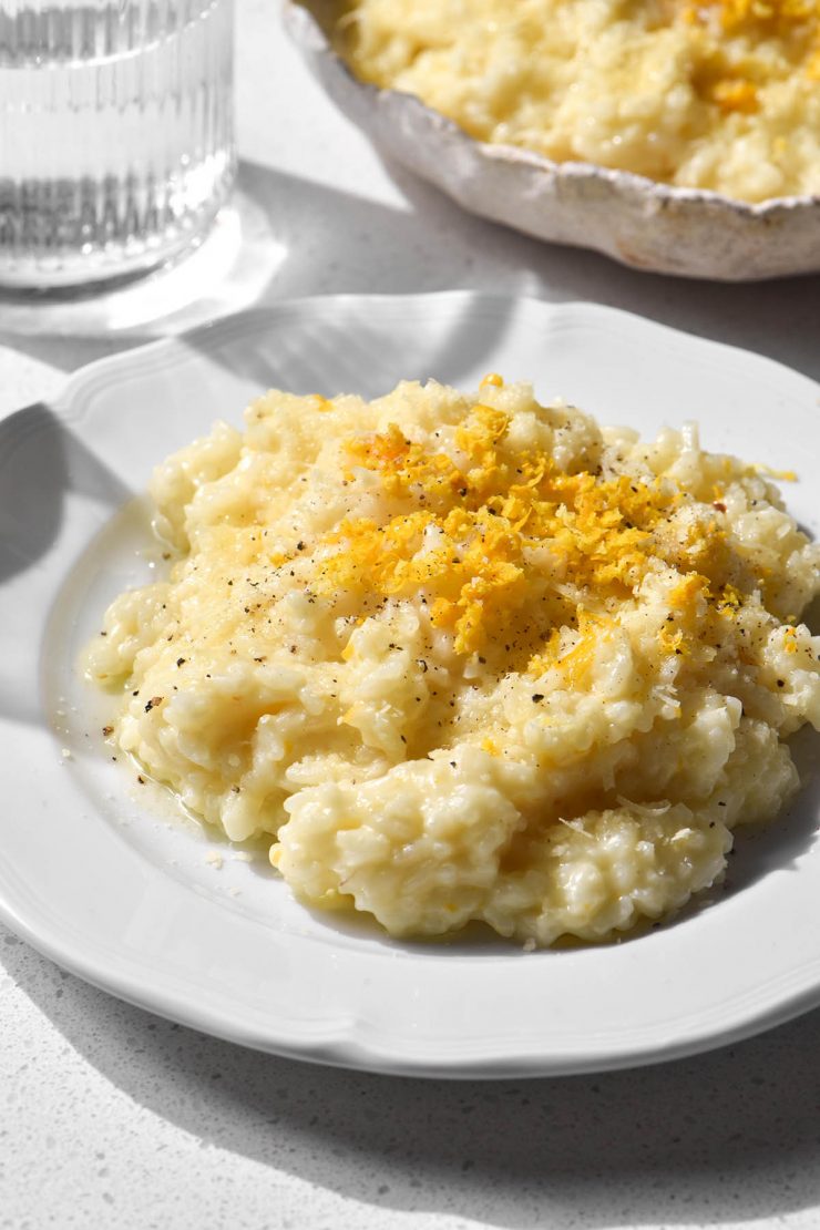 A brightly lit aerial image of two white plates topped with low FODMAP risotto al limone. The plates sit atop a white stone benchtop and two brightly lit glasses of water sit to the left of the plates.