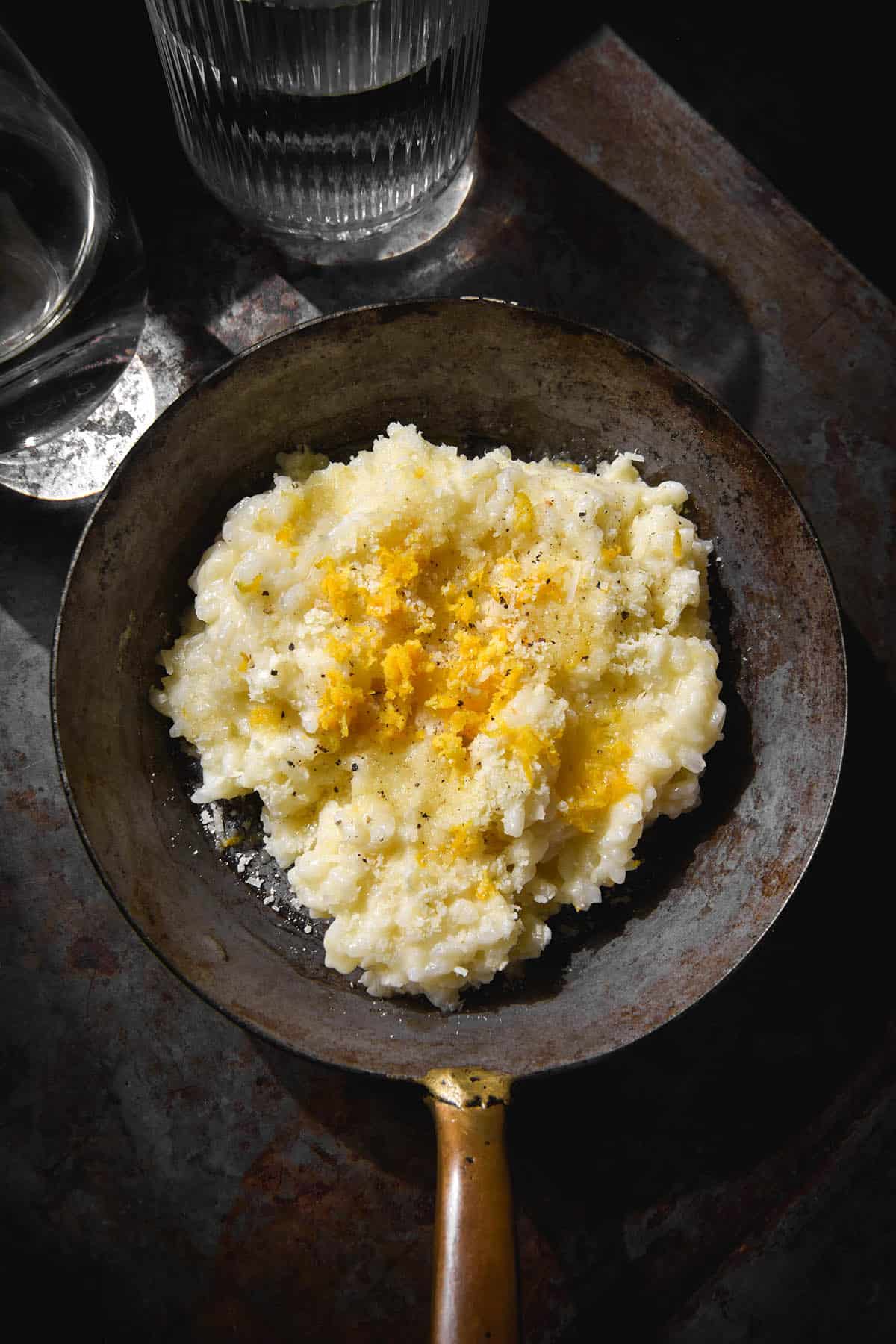 A dark and moody image of a rustic mini skillet of low FODMAP risotto al limone atop a dark grey steel backdrop. Two glasses of water sit to the top left of the image.