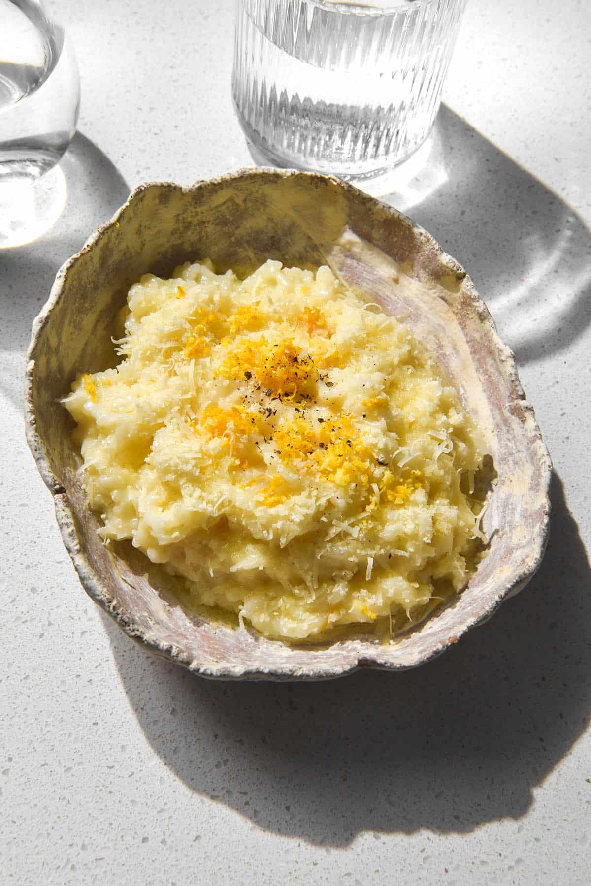 A brightly lit aerial image of a white ceramic bowl filled with low FODMAP risotto al limone. The risotto is topped with garlic infused oil, grated parmesan, lemon zest and black pepper. It sits atop a white stone bench top and a sunlit glass of water sits to the top right of the image.