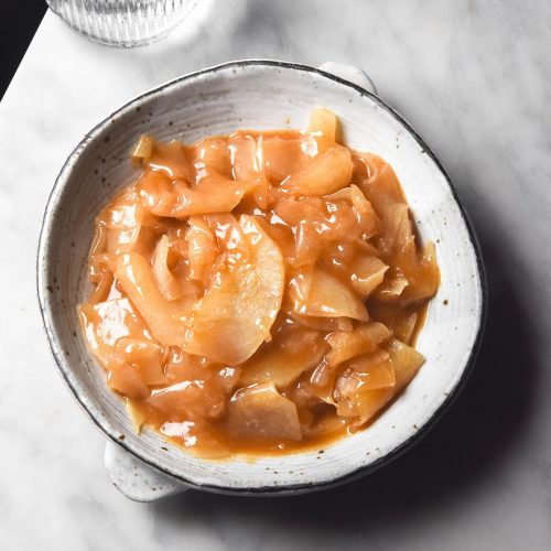 An aerial image of a white speckled ceramic bowl filled with low FODMAP stewed apple. The bowl sits on a white marble table and two sunlit water glasses sit in the top left of the image