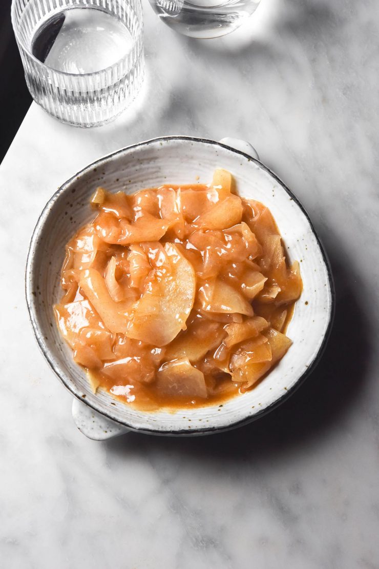 An aerial image of a white speckled ceramic bowl filled with low FODMAP stewed apple. The bowl sits on a white marble table and two sunlit water glasses sit in the top left of the image