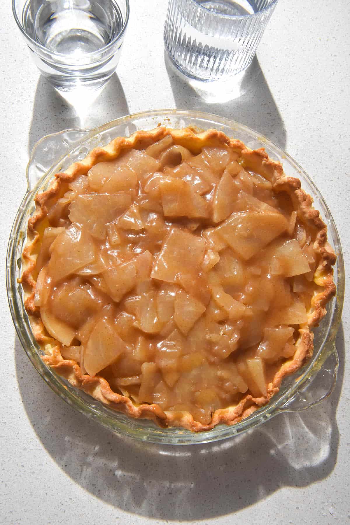 An aerial image of a gluten free apple pie crust in a glass pie plate being filled with low FODMAP stewed apple. The pie plate sits atop a white stone bench top and two sunlit glasses of water sit to the top of the image.