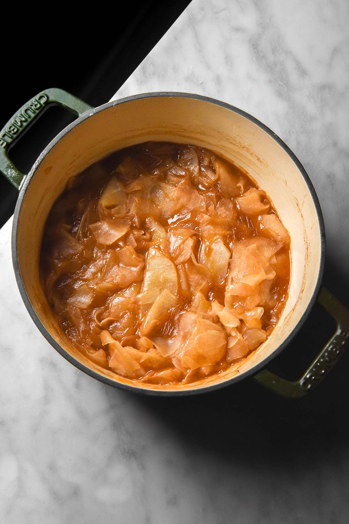 A moody aerial image of a big pot filled with low FODMAP stewed apple made with jicama. The pot sits on a white marble table with a black backdrop in the background. 